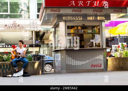 Mads Jacobsen, Straßenmusiker, Gitarre gespielt, sitzt an der Wand neben dem Hotdog-Stand in der Rotenturmstraße in Wien, während ein Paar Mittagessen kauft Stockfoto