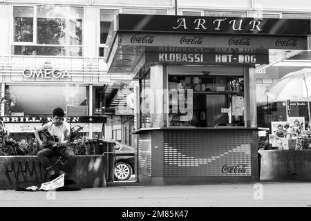 Mads Jacobsen, Straßenmusiker, Gitarre gespielt, sitzt an der Wand neben dem Hotdog-Stand in der Rotenturmstraße in Wien, während ein Paar Mittagessen kauft Stockfoto