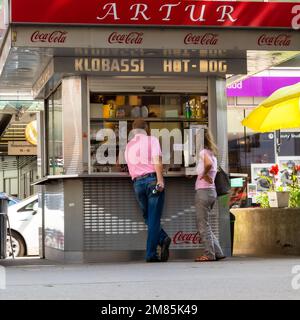 Mads Jacobsen, Straßenmusiker, Gitarre gespielt, sitzt an der Wand neben dem Hotdog-Stand in der Rotenturmstraße in Wien, während ein Paar Mittagessen kauft Stockfoto