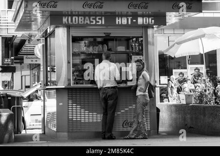 Mads Jacobsen, Straßenmusiker, Gitarre gespielt, sitzt an der Wand neben dem Hotdog-Stand in der Rotenturmstraße in Wien, während ein Paar Mittagessen kauft Stockfoto