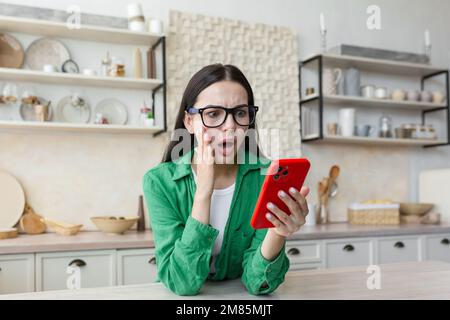 Eine besorgte junge Frau steht zu Hause in der Küche und hält das Telefon in der Hand. Schockiert, dass sie die Nachrichten liest, eine Nachricht erhielt, ihr Konto überprüft. Stockfoto