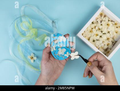Frau verziert Sperrholzrohling in Form eines Ostereiers mit gepressten getrockneten Blumen. Trendige Frühlingsfarben. Künstlerische Ideen für Ostern. Stockfoto