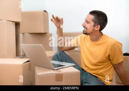 Ein glücklicher Weißer mit den Schlüsseln seines neuen Wohnhauses und einem Laptop zwischen den Kisten. Stockfoto
