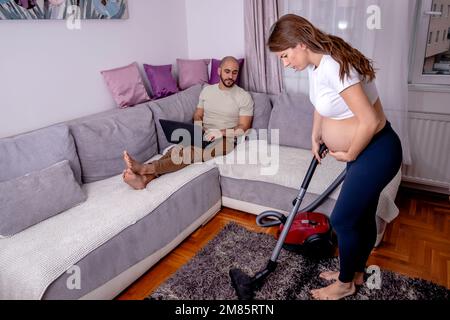 Eine müde schwangere Frau, die Schmerzen hat, während sie ihr Haus putzt. Die Frau saugt das Wohnzimmer, während ihr Mann sich auf dem Sofa entspannt. Stockfoto