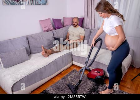 Eine müde schwangere Frau, die Schmerzen hat, während sie ihr Haus putzt. Die Frau saugt das Wohnzimmer, während ihr Mann sich auf dem Sofa entspannt. Stockfoto