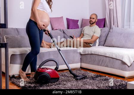 Eine müde schwangere Frau, die Schmerzen hat, während sie ihr Haus putzt. Die Frau saugt das Wohnzimmer, während ihr Mann sich auf dem Sofa entspannt. Stockfoto