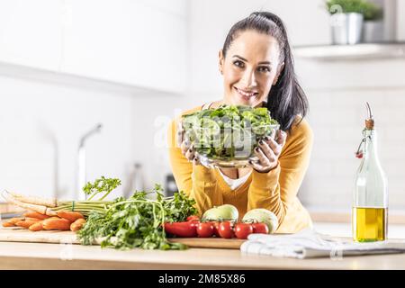 Lächelnde Frau mit einer Schüssel voller Spinatblätter zusammen mit frischem Gemüse und Olivenöl in ihrer Küche. Stockfoto
