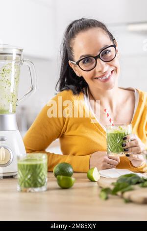 Glückliche Frau mit Spinat-Smoothie-Getränk oder vegetarischem Shake, die in ihrer Küche sitzt. Stockfoto