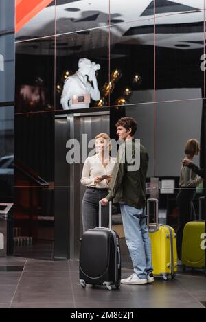 Lächelndes Paar mit Koffer, die wegschauen, während man mit Reisetaschen in einem modernen Hotel steht, Stockbild Stockfoto
