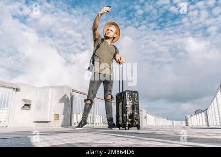 Ganzkörperportrait eines Hipster-Mannes, der ein Selfie gemacht hat Mit einem Koffer auf den Straßen der Stadt Stockfoto