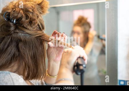 Reife Frau bügelt ihr Haar vor dem Spiegel im Badezimmer ihrer Wohnung. Stockfoto