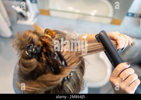 Blonde Frau bügelt ihr Haar mit einem elektrischen Gerät im Badezimmer, Draufsicht. Stockfoto