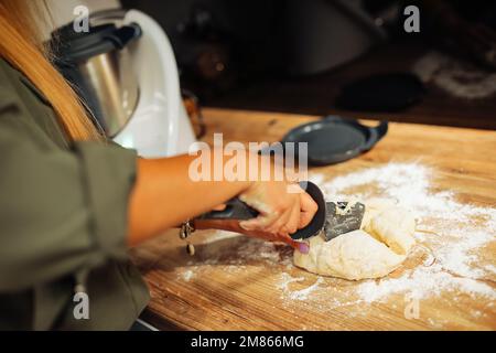 Frauenhände, die Knete mit dem Messer in Stücke schneiden, selektiver Fokus. Kochen Sie in der Küche zu Hause auf einer hölzernen Arbeitsfläche. Stockfoto