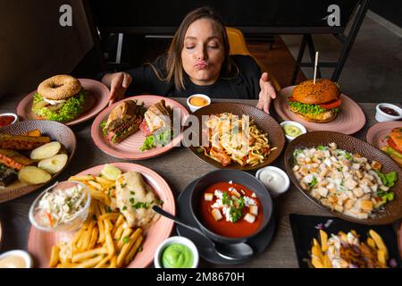 Hungrige Frau mit Vergnügen riecht nach köstlichem Essen auf Tellern, die den Tisch füllen. Pommes Frites, Suppe, Salat, Burger, Saucen zum festlichen Abendessen. Essen, viele Stockfoto