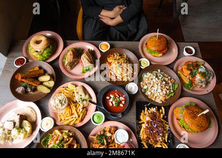 Foto von oben von einer Frau, die an einem Tisch mit köstlichem Essen auf Tellern sitzt. Pommes Frites, Suppe, Salat, Burger, Saucen zum festlichen Abendessen. Essen, viele Gerichte Stockfoto