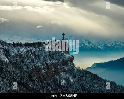 Luftaufnahmen mit einem Kirchenemblem auf einem schneebedeckten Berg, umgeben von Kiefern, alles unter einem bewölkten Himmel in der französischen Landschaft Stockfoto