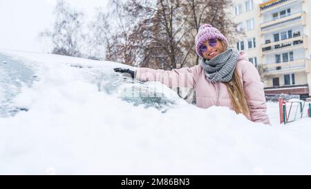 Junge lächelnde Frau, die den ersten Schnee von der Windschutzscheibe ihres Autos säubert. Hochwertiges Foto Stockfoto