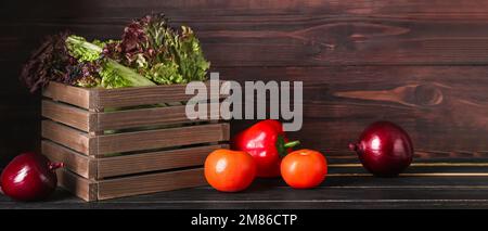 Box mit frischem Lollo Rosso Salat und Gemüse auf dunklem Holzhintergrund Stockfoto