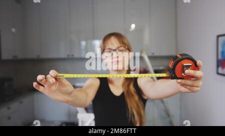 Mittellange Aufnahme einer langhaarigen Frau mit Brille, die ein offenes Maßband hält, Werkzeuge zur Renovierung im Hintergrund. Hochwertiges Foto Stockfoto
