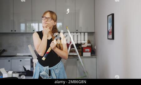 Blonde Frau mit einem Hammer in der Hand, die über die Renovierung in der Küche nachdenkt, Werkzeuge im Hintergrund. Hochwertiges Foto Stockfoto