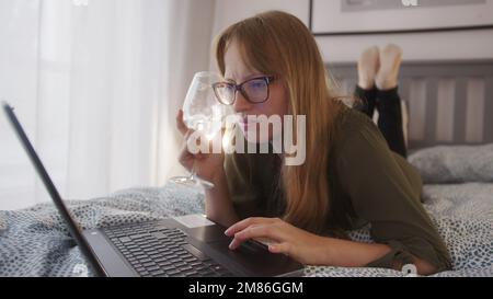 Rothaarige Frau, die ihren Laptop im Bett benutzt und Weißwein trinkt. Hochwertiges Foto Stockfoto