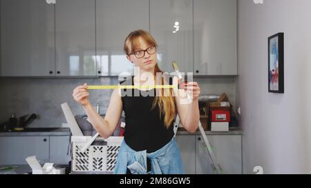 Zeit für die Renovierung, junge Frau in schwarzem ärmellosem Hemd mit langem blondem Haar und Brille, die ein Maßband in der Küche hält. Hochwertiges Foto Stockfoto