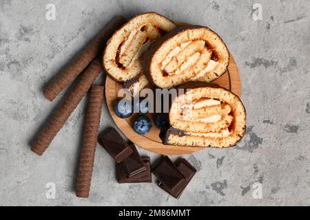 Schneidebrett mit leckeren Kuchenrollen, Schokolade, Waffelrollen und Blaubeeren auf Grunge-Hintergrund Stockfoto