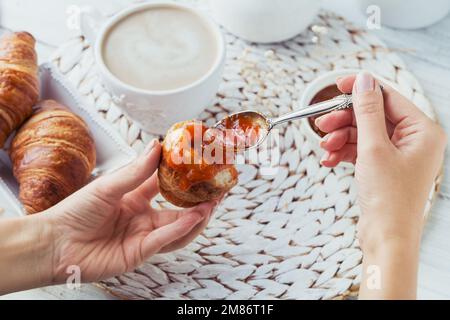 Köstliches Frühstück mit frischen Croissants und einer Tasse Kaffee serviert mit Marmelade auf weißem Holzhintergrund. Frauenhand mit Löffel mit Marmelade. Stockfoto