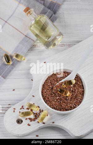 Braune Leinsamen oder Leinsamen in einer kleinen Schüssel und Gelatinekapseln mit Omegaöl auf weißem Holzhintergrund. Stockfoto