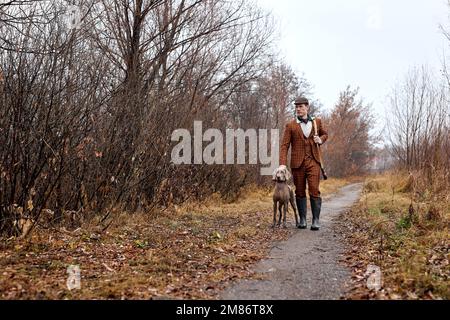 Selbstbewusster männlicher Jäger mit nettem Jagdhund Weimaraner, Porträt in ländlicher Gegend während der Jagdsaison, hübscher weißer Kerl in braunem Anzug, zu Fuß zu erreichen Stockfoto