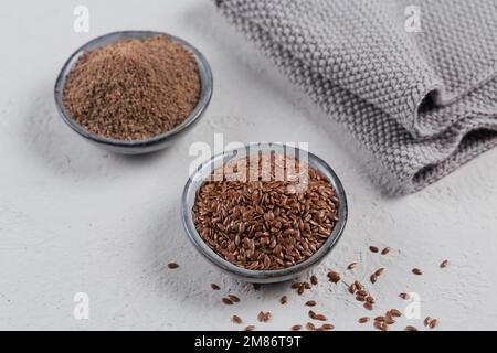 Braune Leinsamen und gemahlene oder zerkleinerte Leinsamen oder Leinsamen in einer kleinen Schüssel auf hellgrauem Hintergrund. Stockfoto