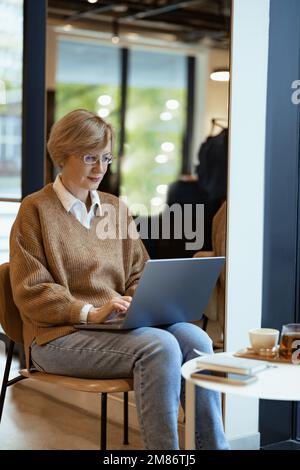 Fokussierte, reife Geschäftsfrau mittleren Alters, die in einem gemütlichen Café mit einem Laptop arbeitet und im Internet surft Stockfoto
