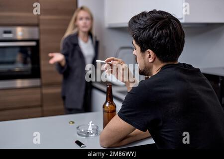 Familienstreit. Unglückliche junge Kaukasierfamilie während Konflikten in der Küche zu Hause. Konzentrieren Sie sich auf den Kerl, der raucht und Alkohol trinkt. Ehepaar streitet sich. Stockfoto