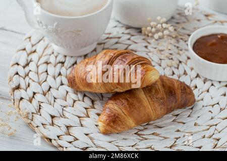 Köstliches Frühstück mit frischen Croissants und einer Tasse Kaffee serviert mit Marmelade auf weißem Holzhintergrund. Köstliches Backen Stockfoto