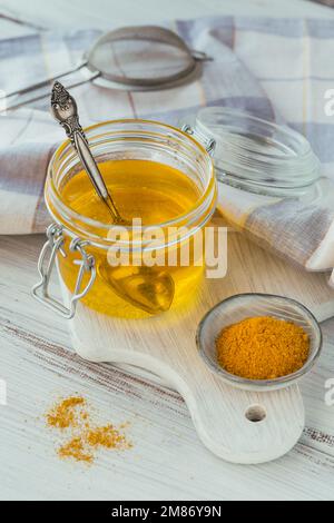 Hausgemachter Ghee oder gereinigte Butter in einem Glas und Kurkuma auf einem weißen Holztisch. Stockfoto