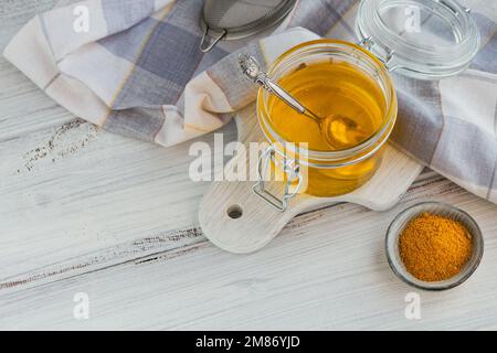 Hausgemachter Ghee oder gereinigte Butter in einem Glas und Kurkuma auf einem weißen Holztisch. Stockfoto