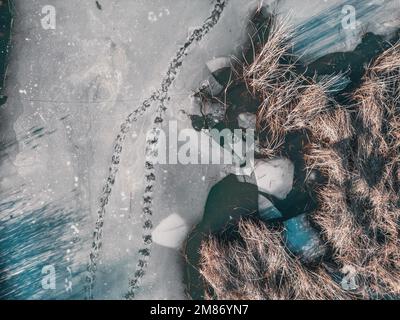 Panoramablick aus der Vogelperspektive auf einen gefrorenen See mit Tierspuren und gesprungenem Eis. Tierspuren auf der gefrorenen Oberfläche des Sees. Eis am See gebrochen Stockfoto