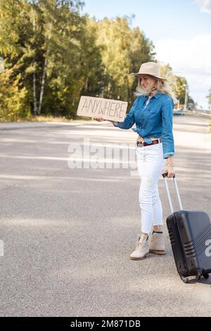 Eine Frau, die im Wind Haare bläst, versucht, das Auto mit einem Pappposter auf einer leeren Autobahn zu stoppen. Frau mit Hut, mit Koffer, Flucht aus der Stadt zum Mitnehmen Stockfoto