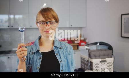 Junge Frau mit Schutzbrille, die einen Schraubenschlüssel nach oben in der Küche hält, Werkzeuge im Hintergrund. Hochwertiges Foto Stockfoto