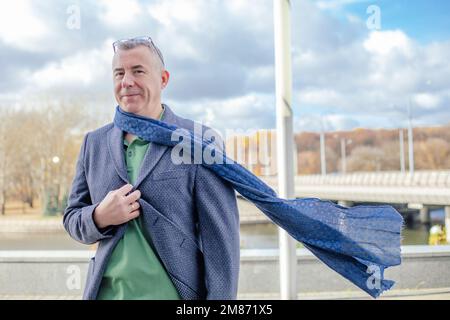 Zufriedener, selbstbewusster, lächelnder, grilliger Geschäftsmann mit Brille auf dem Kopf in warmer Jacke, Schal spazieren, Schauer auf der Straße Stockfoto