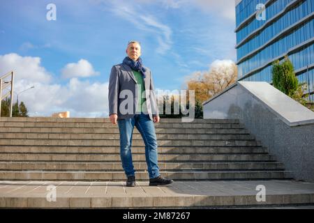 Seriöser, gereifter Geschäftsmann in Jacke und Schal, Kamera, stellen Sie sich auf die Treppe neben dem Bürogebäude. Beförderung Stockfoto