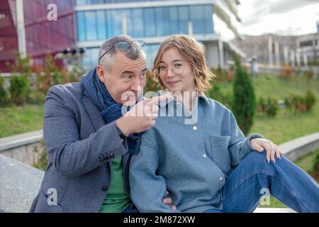 Ein lächelnder, reifer Mann, der einer jungen Frau etwas erklärt und zeigt, sitzt auf dem Geländer gegen das Universitätsgebäude. Familie Stockfoto