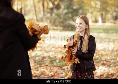 Sehen Sie ein lächelndes, glückliches, sorgenfreies blondes Mädchen in schwarzen Jacken und sammeln Sie Blätter im goldenen Herbstwald Stockfoto