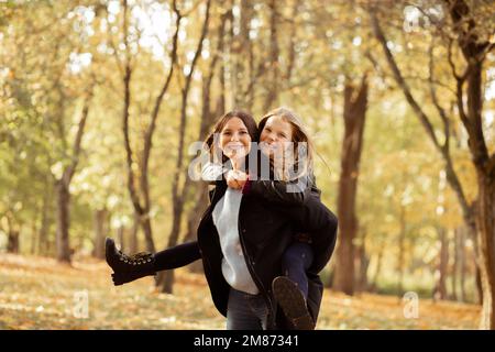 Fröhliche, verspielte, positive, lächelnde Familie der mutter hält die Tochter auf dem Rücken und läuft am Wochenende im goldenen Herbstwald Stockfoto