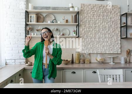Fröhliche und glückliche junge Frau, Hausfrau, Studentin singt in der Küche als Mikrofon in einen Küchengegenstand und tanzt. Kocht gerade. Stockfoto