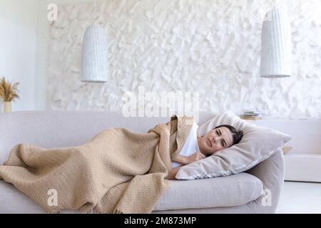 Eine junge, müde Frau liegt zu Hause auf dem Sofa, das mit einer Decke bedeckt ist. Krank, sich schlecht fühlen, Depressionen, harter Tag. Stockfoto