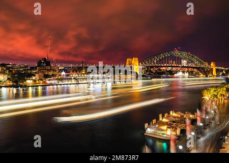 Leichte Wanderwege von Fähren, die am Circular Quay in Sydney, Australien, ankommen und abfahren, unter dem roten Nachglühen des Sonnenuntergangs mit dem Sydney Harbour Br Stockfoto