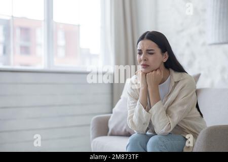 Eine traurige und besorgte junge Frau leidet an Depressionen, psychischen Störungen. Sie sitzt durchdacht im Krankenhauszimmer auf dem Sofa am Fenster und legt ihren Kopf auf ihre Hände. Stockfoto