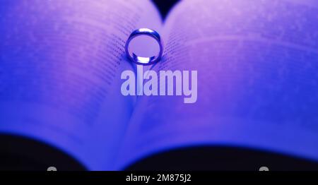 Ehering in offenem Buch und herzförmiger Schatten. Blaues Neonlicht beleuchtet Seiten. Einsatzvorschlag. Konzept des Valentinstages. Webbanner. Stockfoto