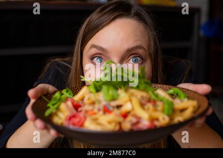 Porträt einer Frau mittleren Alters, die mit breiten Augen in die Kamera starrt und leckere Pasta mit Kirschtomaten, Rucola und Käse zeigt. Stockfoto